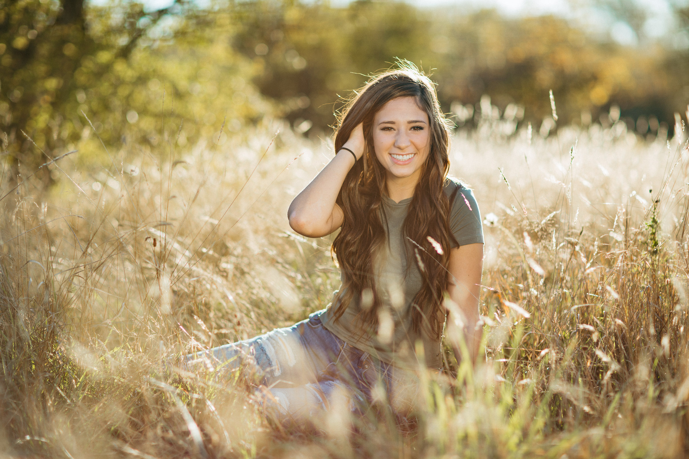 High school senior wearing a white dress sitting in a grassy knoll with late afternoon sun glowing from behind.