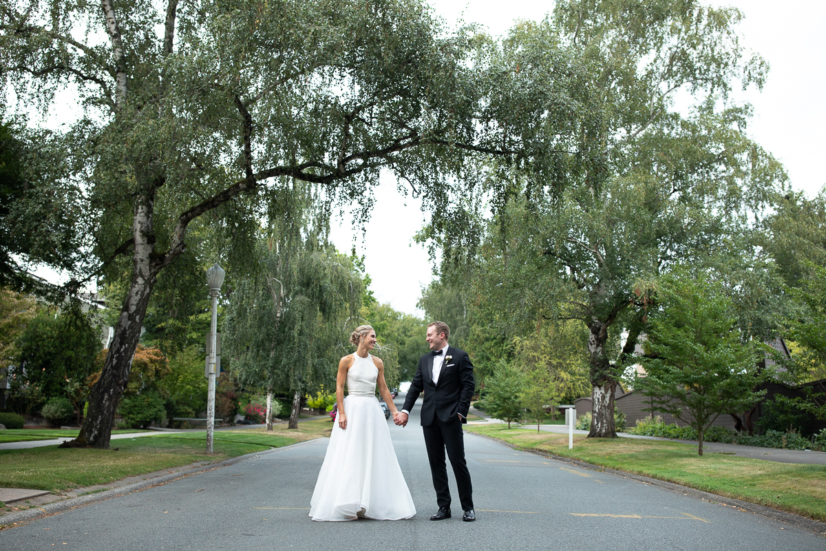 Bride and groom holding hands on tree-lined street in front of Seattle Yacht Club