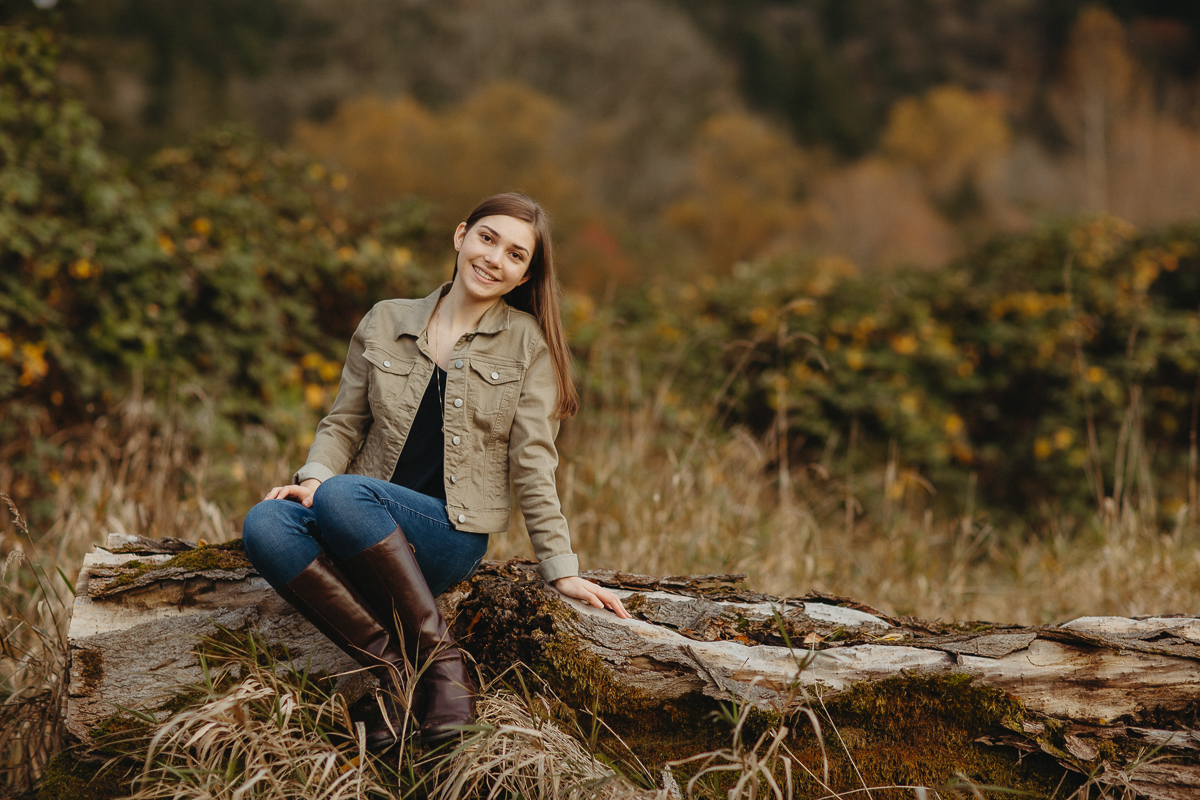 Young woman smiling, sitting on log, fall colors backdrop.